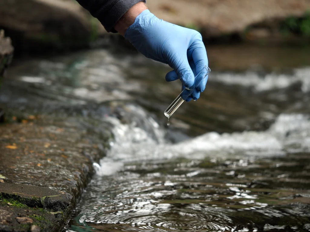 A professional with a gloved hand is using a water collecting tube to gather water quality data.